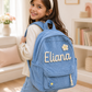 Girl wearing a blue backpack with 'Eliana' embroidered on it in a cozy room.